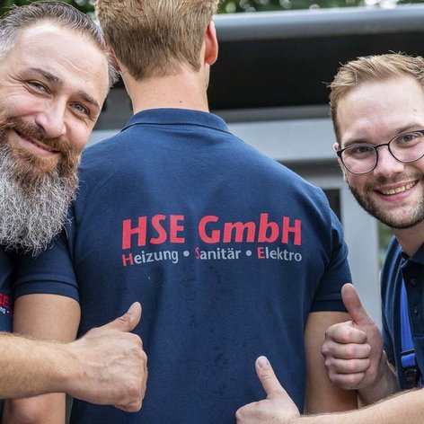 Die beiden Männer posieren fröhlich, einer zeigt auf das Logo 'HSE GmbH' auf dem Rücken des anderen.