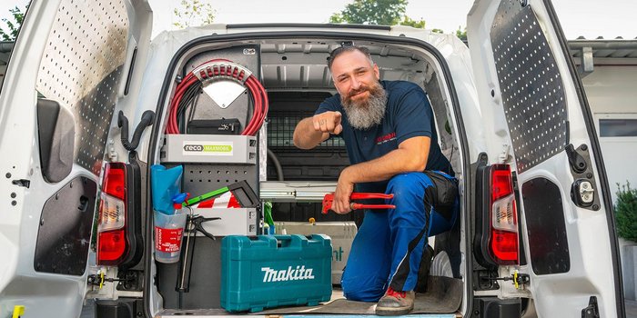 Der Mann im blauen Arbeitshemd posiert lächelnd in einem offenen Lieferwagen, umgeben von verschiedenen Werkzeugen und Materialien.