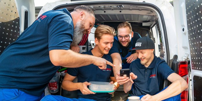 Vier Männer in blauen Arbeitshemden sitzen in einem Lieferwagen und schauen gemeinsam auf ein Smartphone, lächelnd und interessiert.