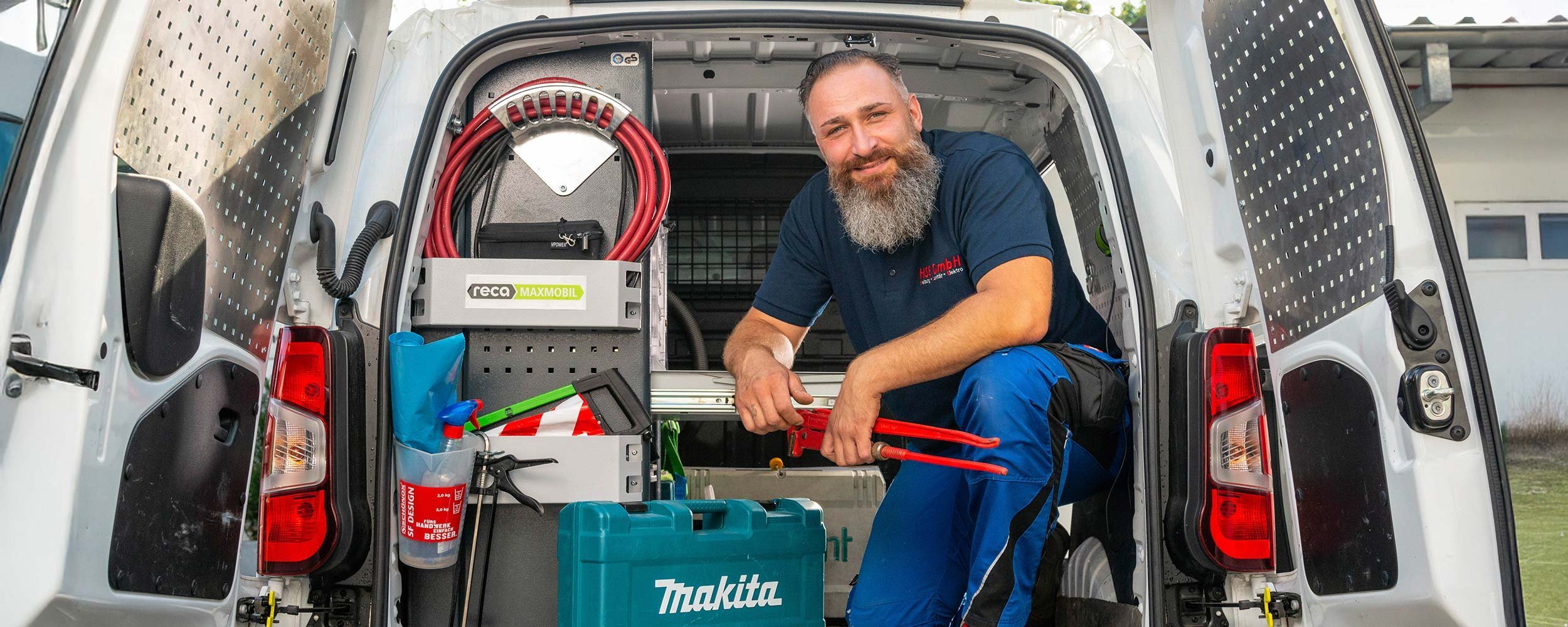 Ein Handwerker mit Bart sitzt in einem Lieferwagen, umgeben von Werkzeugen und Ausrüstung, und lächelt freundlich.