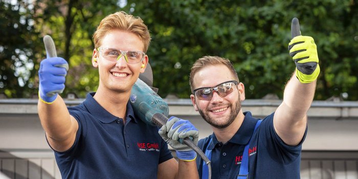 Zwei Handwerker lächeln und zeigen Daumen hoch, während sie eine elektrische Bohrmaschine halten, umgeben von grüner Natur.