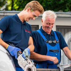 Zwei Männer in Arbeitskleidung lächeln, während sie gemeinsam an einem Werkzeug arbeiten, umgeben von einem hellen, grünen Hintergrund.