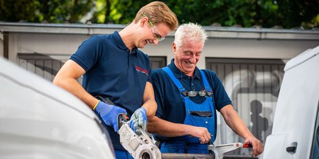 Zwei Männer in Arbeitskleidung lächeln, während sie gemeinsam an einem Werkzeug arbeiten, umgeben von einem hellen, grünen Hintergrund.
