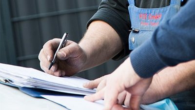 Ein Arbeiter notiert Informationen auf einem Dokument, während eine andere Person ihm dabei zusieht und die Hände auf dem Tisch hat.