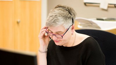 Eine Frau mit grauen Haaren trägt ein Headset und spricht am Telefon, während sie auf ihren Computerbildschirm schaut.