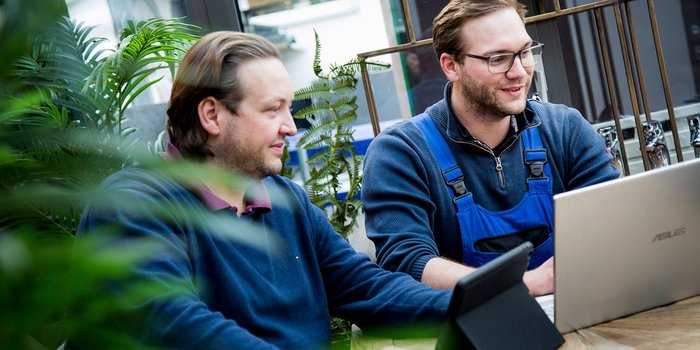 Zwei Männer sitzen an einem Tisch, umgeben von Pflanzen, und arbeiten konzentriert an einem Laptop.