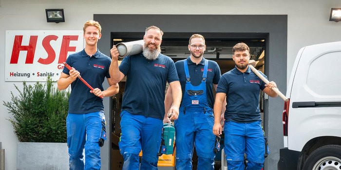 Vier Handwerker in blauen Overalls stehen vor einem Firmengebäude, lächeln und halten Werkzeuge in den Händen.
