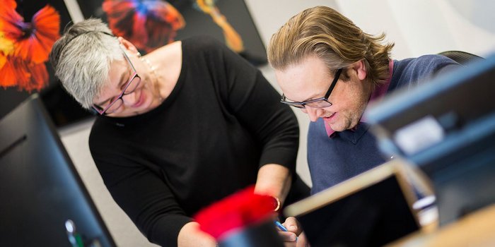 Eine Frau mit grauen Haaren und Brille lächelt, während sie einem Mann mit Brille bei der Arbeit hilft.