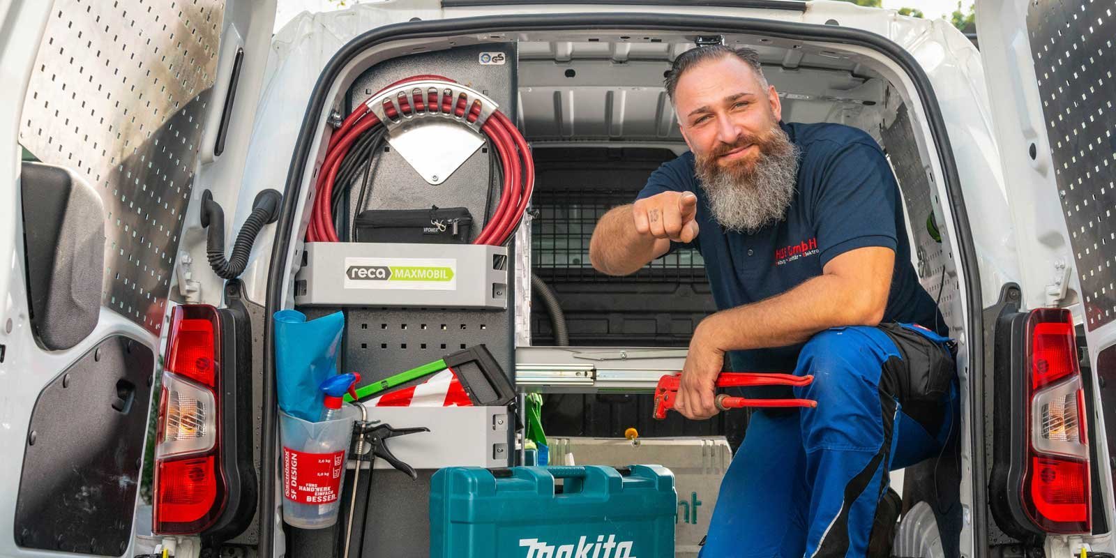 Ein Handwerker mit Bart sitzt in einem Lieferwagen und zeigt mit dem Finger auf die Kamera, um seine Arbeit zu betonen.