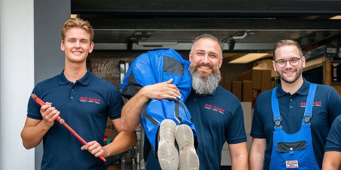 Die Gruppe von drei Handwerkern posiert fröhlich vor einer Garage, einer hält einen blauen Anzug, während die anderen lächeln.