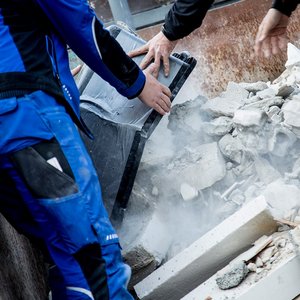 Zwei Arbeiter heben eine schwarze Kiste aus einem Trümmerhaufen, während Staub aufwirbelt und die Umgebung chaotisch wirkt.